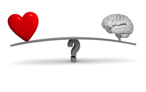 A bright red heart and gray brain sit on opposite ends of a dark gray board balanced on a gray question mark. Isolated on white.